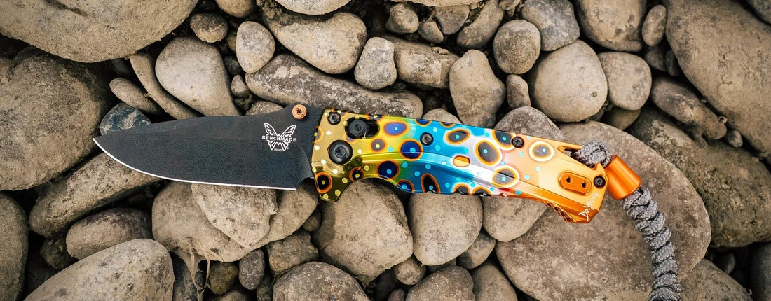 Benchmade folding knife with vibrant peacock feather handle by Derek DeYoung, resting on smooth gray river stones.