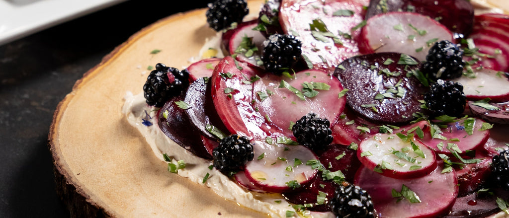 Thinly sliced beet carpaccio on a wooden board, garnished with fresh blackberries and herbs for an elegant presentation.