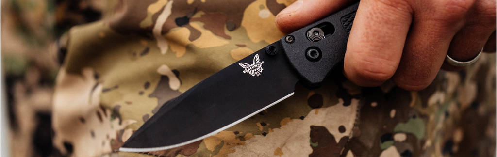 Hand gripping a black Benchmade folding knife with logo, set against camouflage fabric.