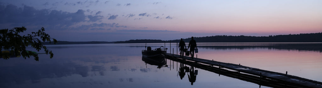 Two people walk a dock at sunset, carrying bags toward a boat on a calm lake near Bowen Lodge.