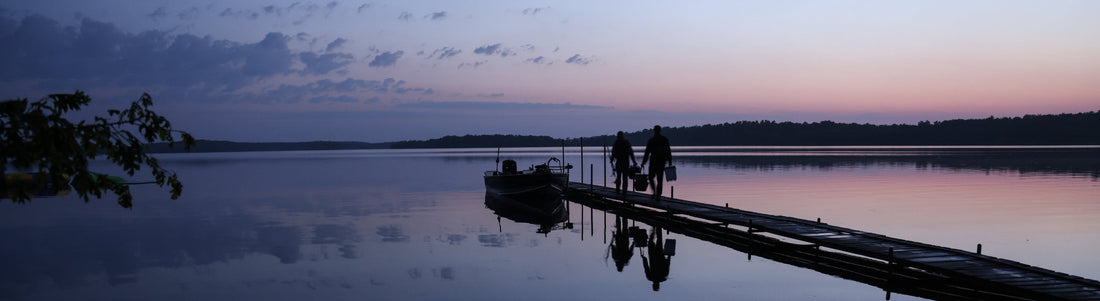 Two people walk a dock at sunset, carrying bags toward a boat on a calm lake near Bowen Lodge.