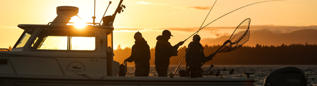 Silhouetted anglers on a boat at sunset, one landing a fish with a net.