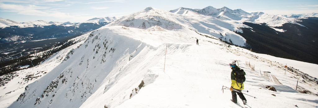 Hiker in a yellow jacket and backpack traverses a snowy mountain ridge, demonstrating essential gear for cold weather carry.