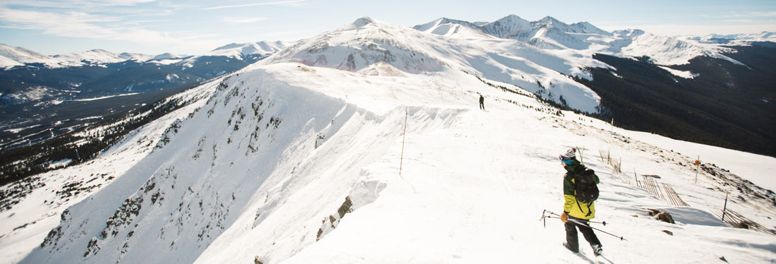 Hiker in a yellow jacket and backpack traverses a snowy mountain ridge, demonstrating essential gear for cold weather carry.