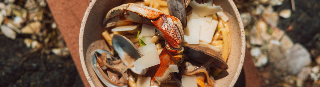 Rustic bowl of strozzapreti with crab claw, mussels, and shaved cheese in bright natural light.