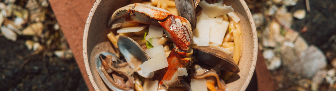 Rustic bowl of strozzapreti with crab claw, mussels, and shaved cheese in bright natural light.