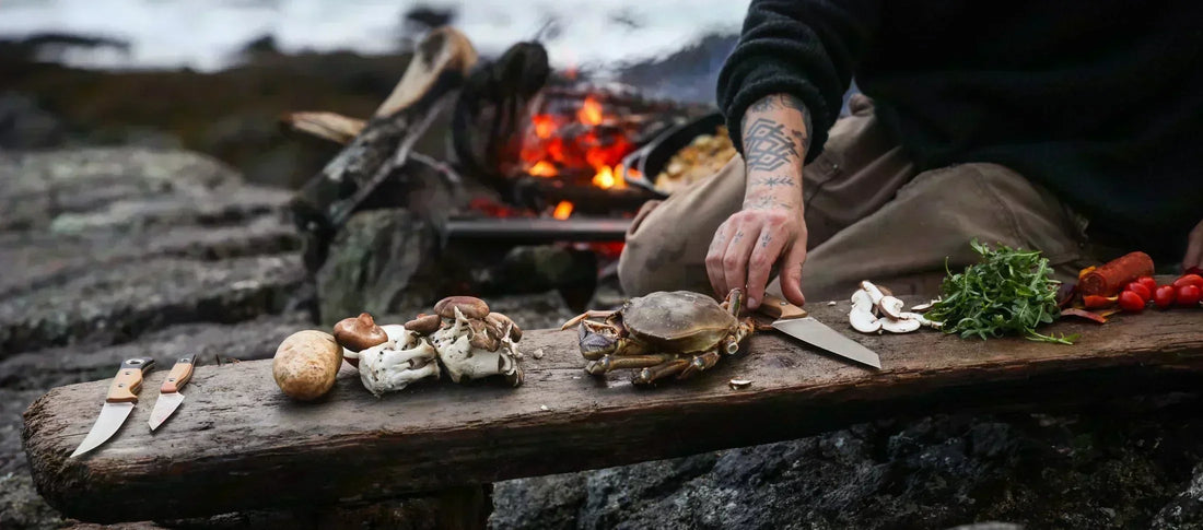 Tattooed hands prep a crab and mushrooms on a rustic wooden plank by a campfire, showcasing sharp outdoor kitchen knives.