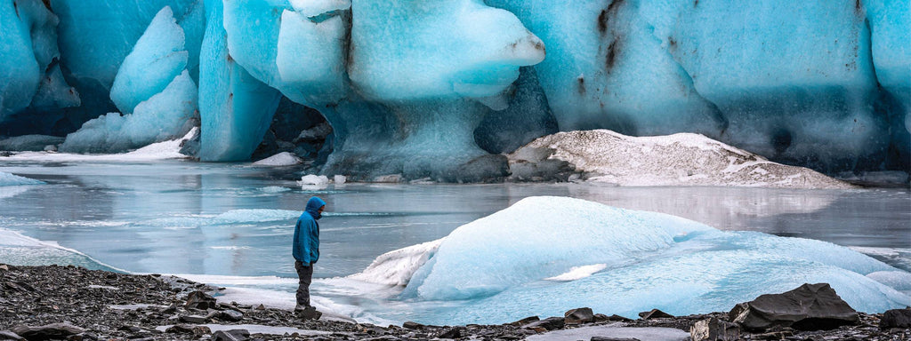 A lone hiker in a blue jacket gazes at towering, vibrant turquoise glaciers and icy waters.