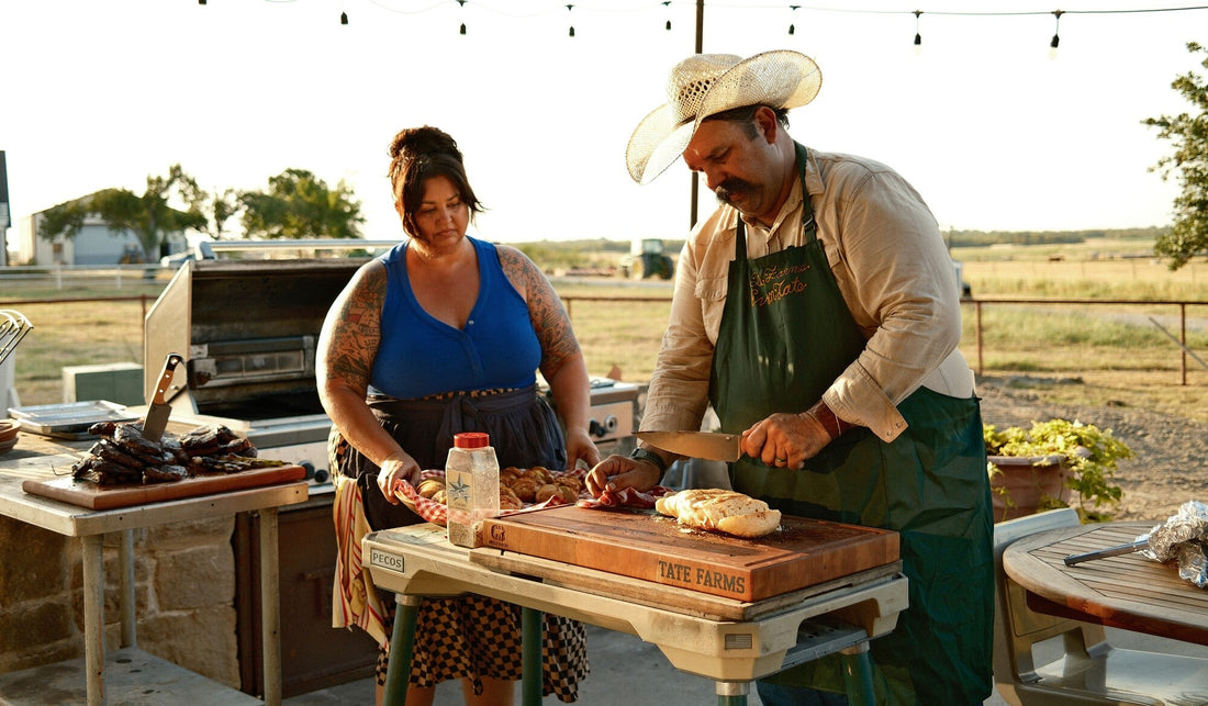 Two people prepare a rustic outdoor barbecue, slicing meat on a wooden board to honor family traditions and legacy.