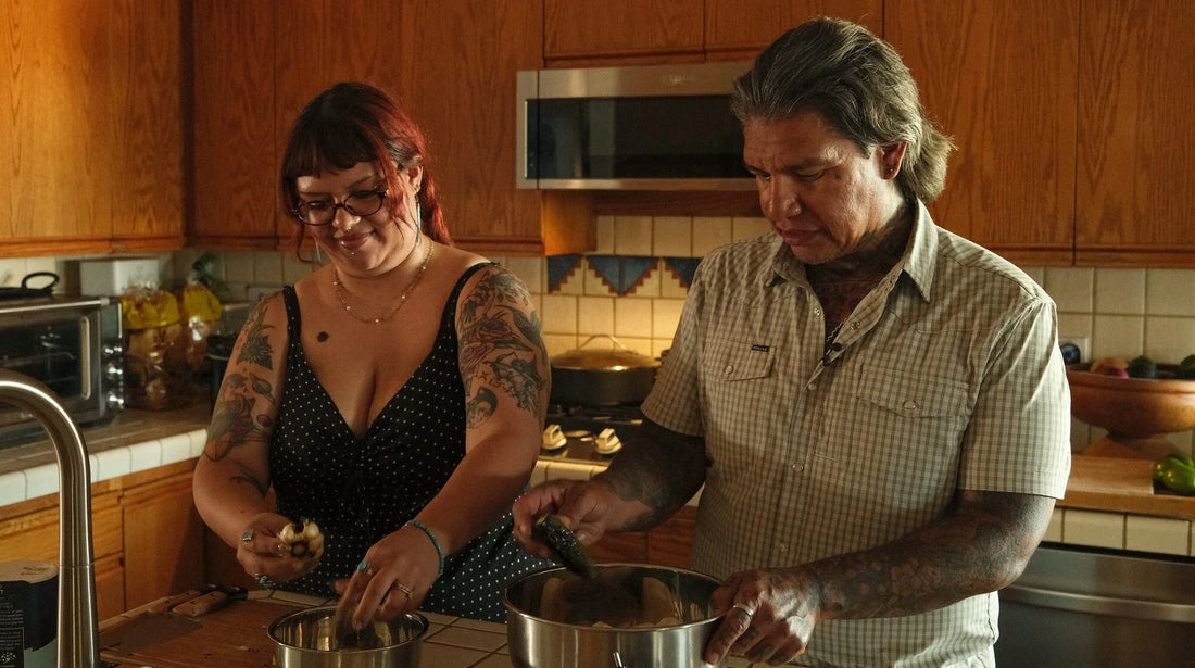 Tattooed chef Daniel Castillo and a smiling woman prepare ingredients together in a warm, wood-paneled home kitchen.