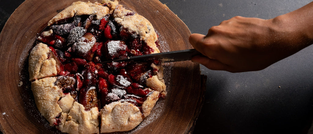 A hand slices a rustic, powdered-sugar-dusted fruit galette on a wooden board, revealing juicy red berries inside.
