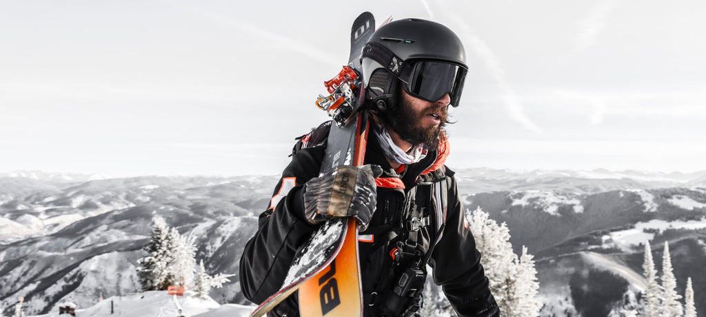 Bearded skier in helmet and goggles carries skis across a snowy mountain ridge, gazing out at the vast winter highlands.