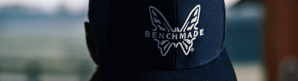 Close-up of a dark blue cap featuring an embroidered white Benchmade butterfly logo against a blurred outdoor background.