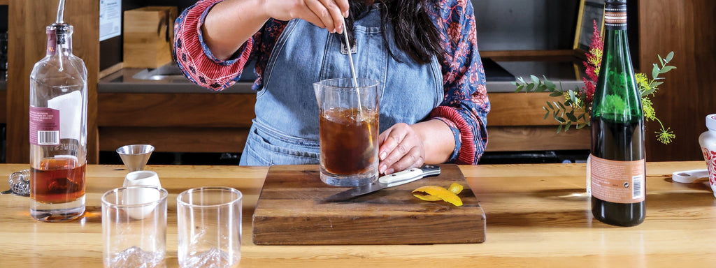 Bartender stirs a rich, amber cocktail on a wooden board, surrounded by bottles and fresh citrus garnish.