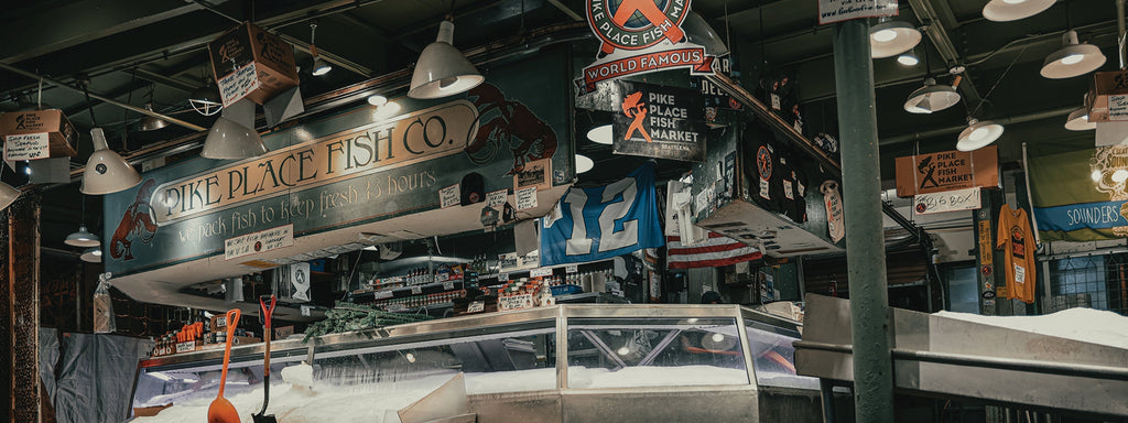 Bustling Pike Place Fish Market stall with hanging signs, ice displays, and a lively, authentic Seattle atmosphere.