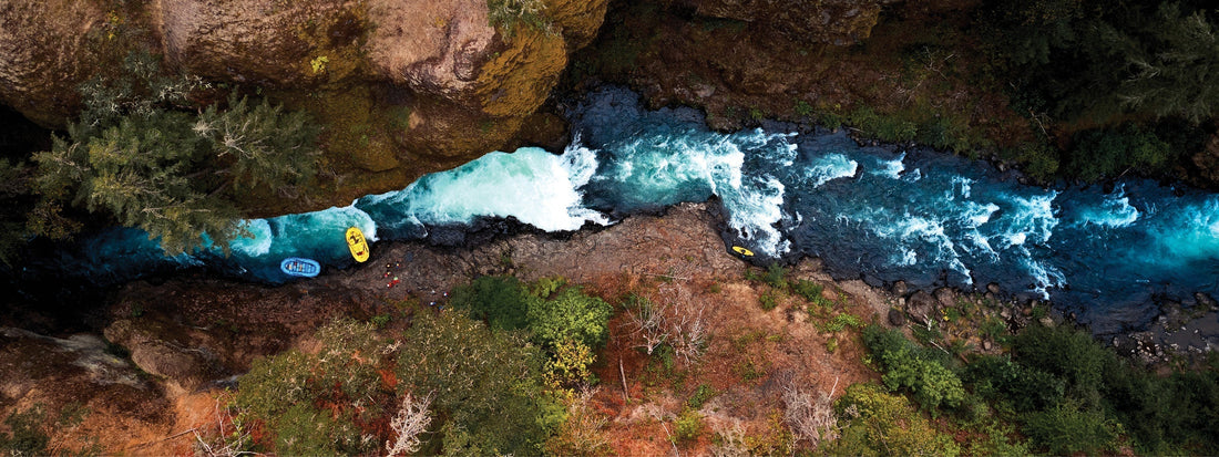 Aerial view of kayakers navigating vibrant turquoise rapids through a rugged, forested canyon.