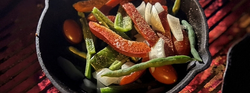 Cast-iron skillet with colorful peppers, onions, and beans over glowing embers for a rustic fajita prep.