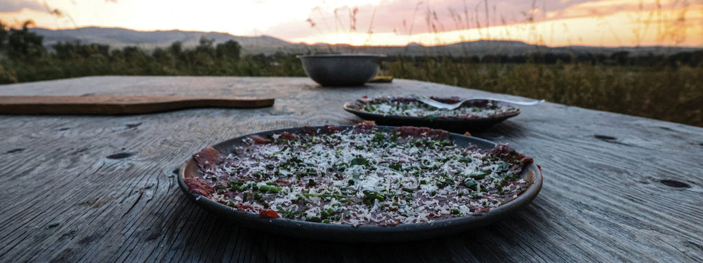 A carpaccio dish on a rustic wooden table at sunset, with fresh herbs and cheese, evoking a serene outdoor dining experience.