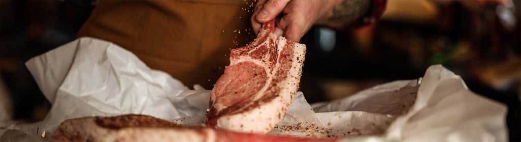 Tattooed hand lifts a raw, spice-rubbed Iberico pork tomahawk from white butcher paper, highlighting rich marbling and texture.