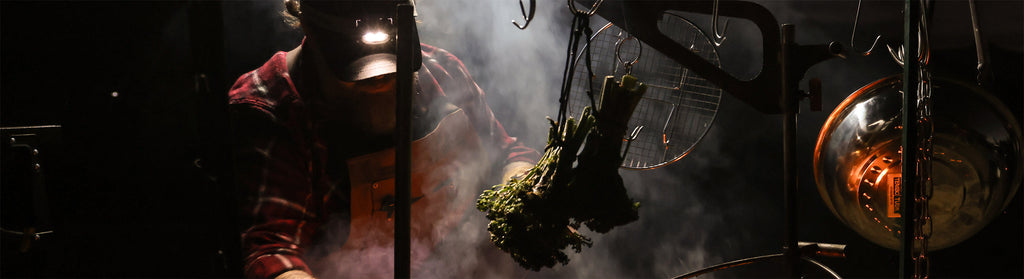 Chef in plaid shirt and headlamp tends to smoky fire-roasted broccolini hanging on a grill.