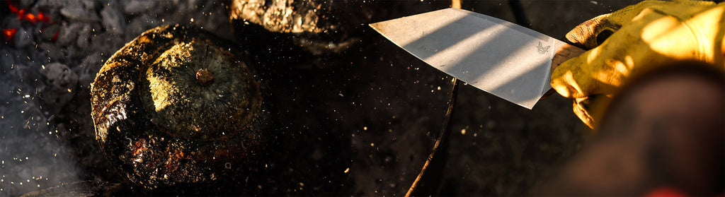 Gloved hand holds a knife near charred squash over glowing embers, capturing the rustic fire-roasting process.