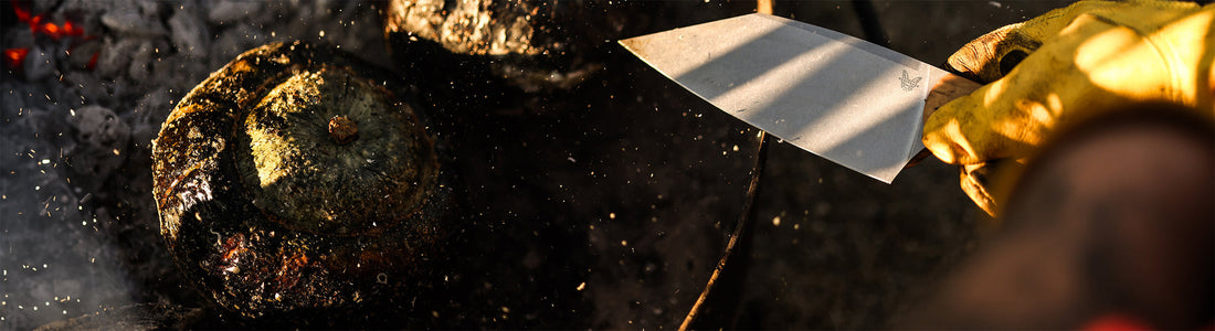 Gloved hand holds a knife near charred squash over glowing embers, capturing the rustic fire-roasting process.