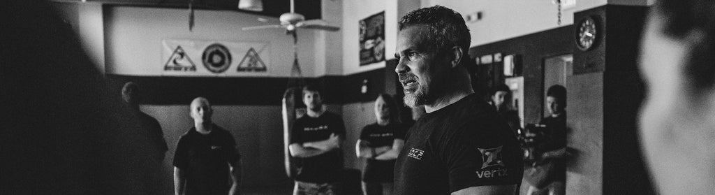 Greg Thompson in a gym, speaking to students. Black-and-white photo capturing the origins of SOCP training.