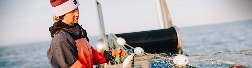 A fisher in a beanie and orange bib handles nets on a boat, bathed in the soft, cool light of the blue hour.