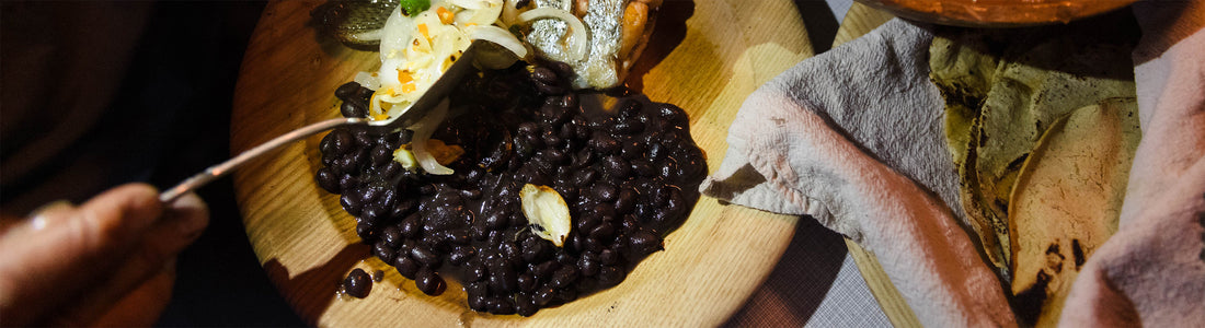 A hand spoons rich, scratch-made black beans from a wooden plate, garnished with onions and herbs.