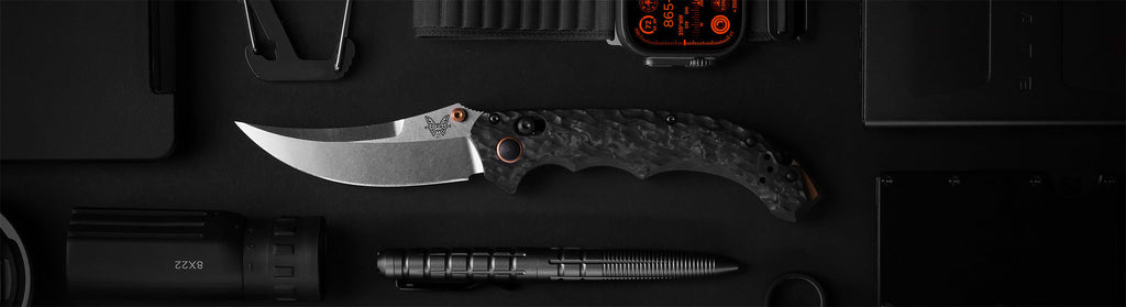 A menacing karambit knife with a carbon fiber handle rests on a dark surface, surrounded by tactical gear.