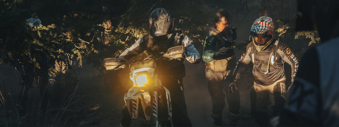 Riders pause in a dusty forest during WLF Enduro Mission 05, headlights glowing through the haze.