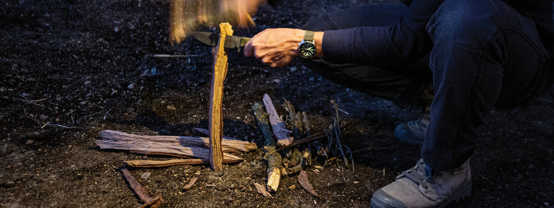 Crouching camper splits a stick with a knife, preparing kindling for a fire in the dim evening light.