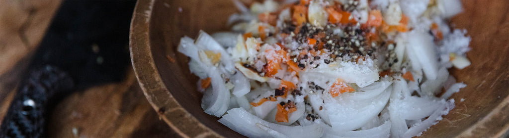 Fresh onions, habanero bits, salt, and pepper in a rustic wooden bowl beside a chef's knife.