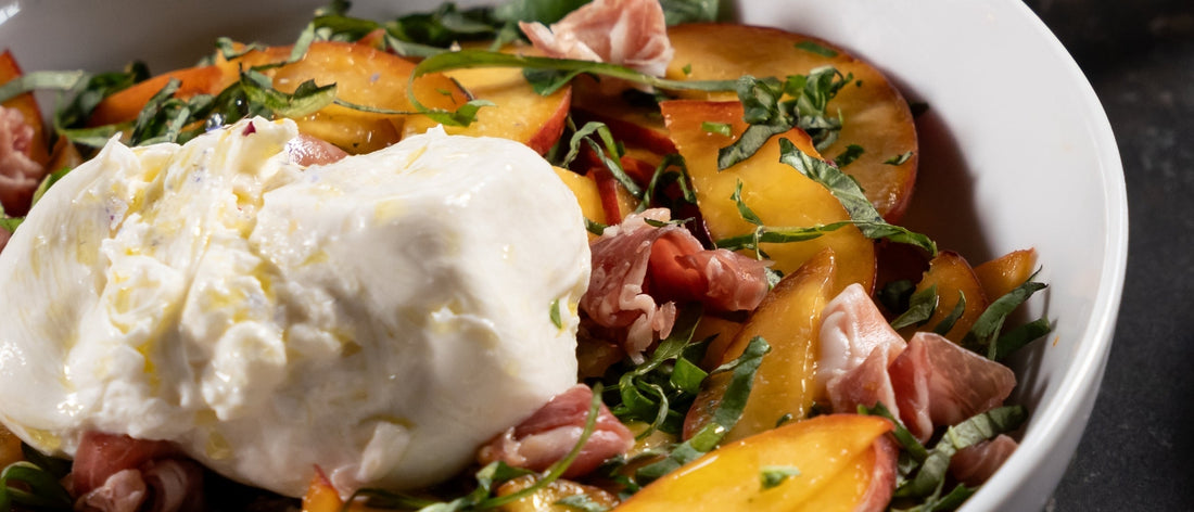 Creamy burrata atop sliced peaches, basil, and prosciutto in a white bowl.