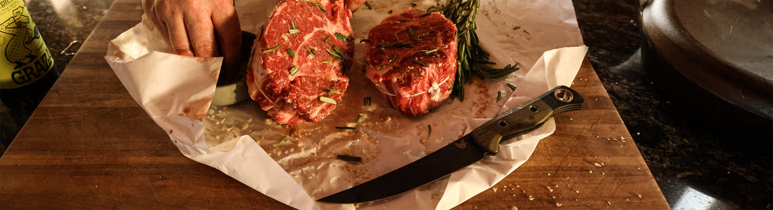 Two raw bone-in steaks garnished with rosemary on butcher paper, with a chef's hand and knife ready for prep.