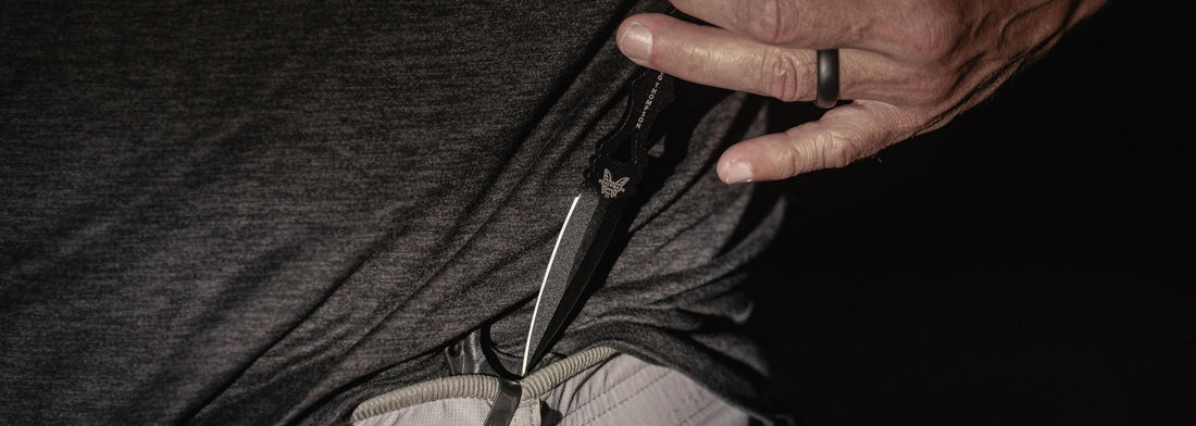 Hand grips a black Benchmade SOCP dagger, its sharp blade emerging from a dark gray shirt in a tactical pose.