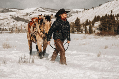 Cowgirl leads saddled horse through snowy field, capturing rugged Western spirit and timeless lifestyle photography.