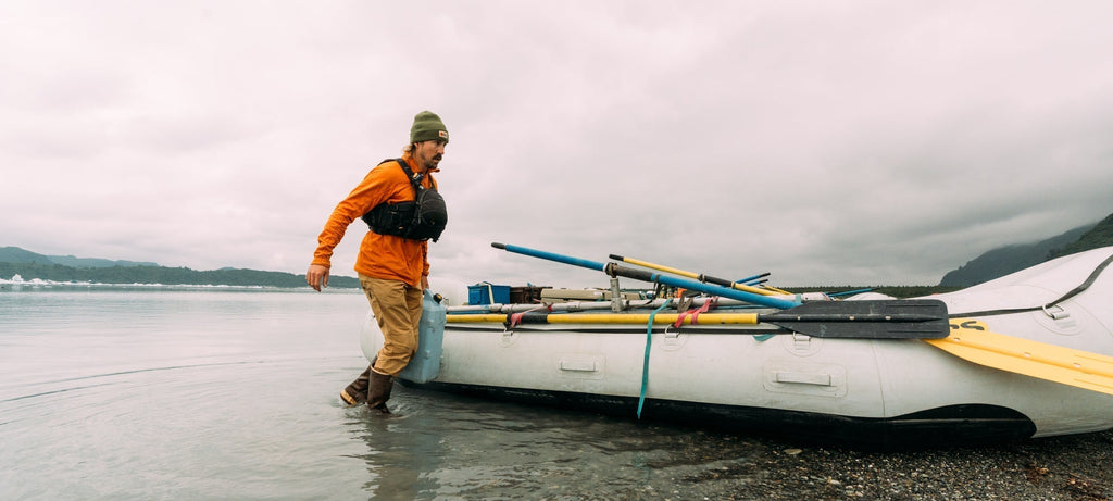 A man in an orange jacket wades into calm waters, pushing a loaded raft under an overcast sky.