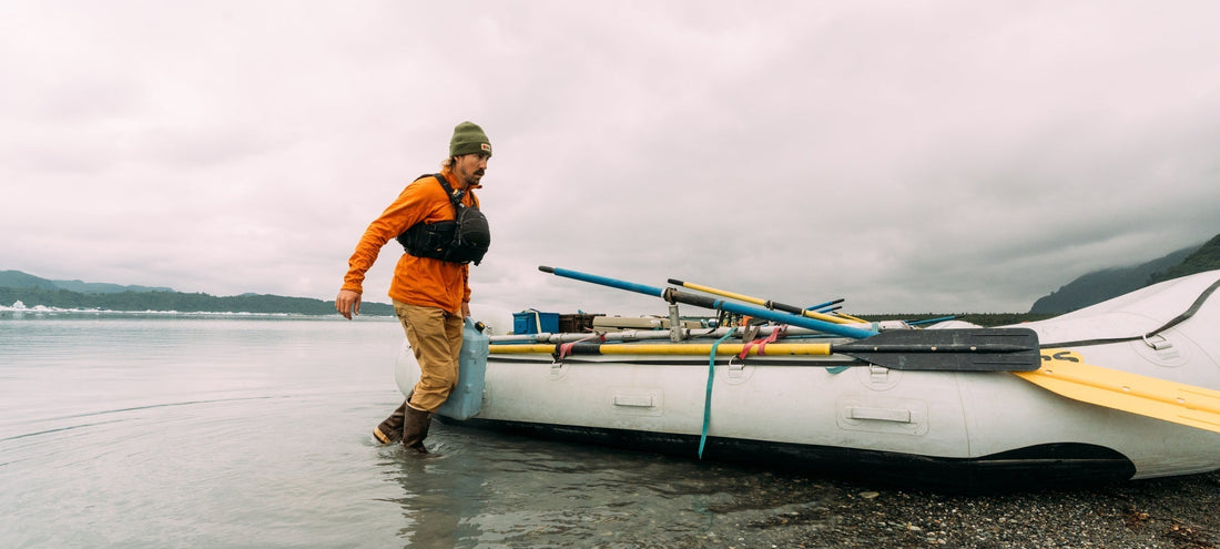 A man in an orange jacket wades into calm waters, pushing a loaded raft under an overcast sky.