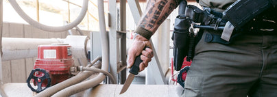 Tattooed hand grips a tactical folding knife near an industrial valve, ready for self-defense in any environment.