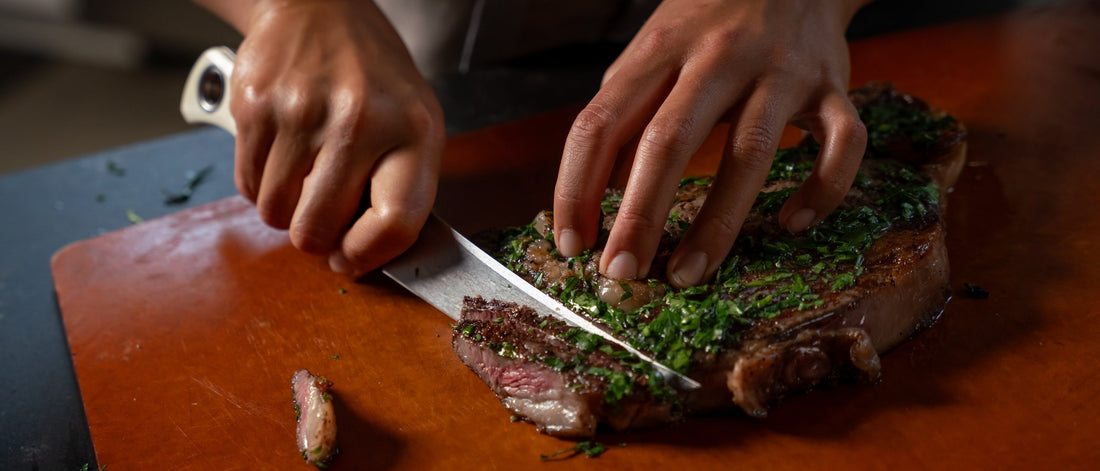 Hands slicing a juicy cast-iron steak topped with fresh chimichurri on a warm wooden board
