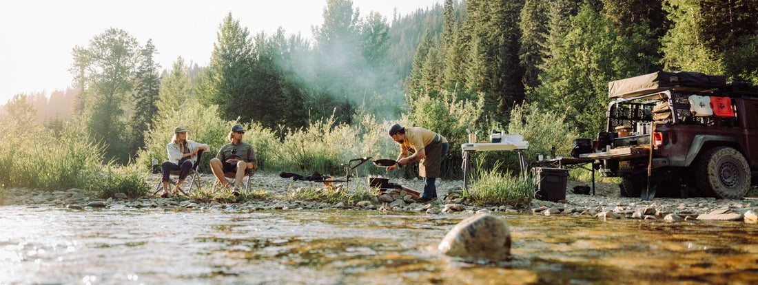 Friends cook over a fire by a river, with a Jeep nearby, embodying the rugged Trail Feast and Benchmade spirit.