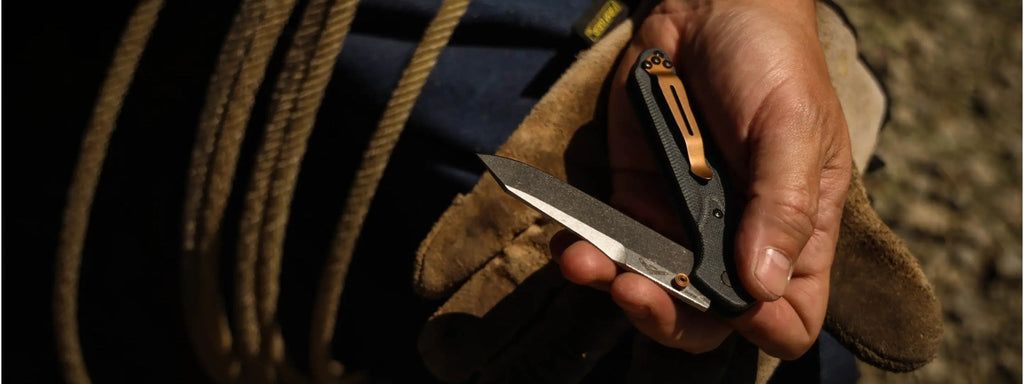 Hand holding a folding knife with a bronze clip, set against rugged rope and leather work gloves.