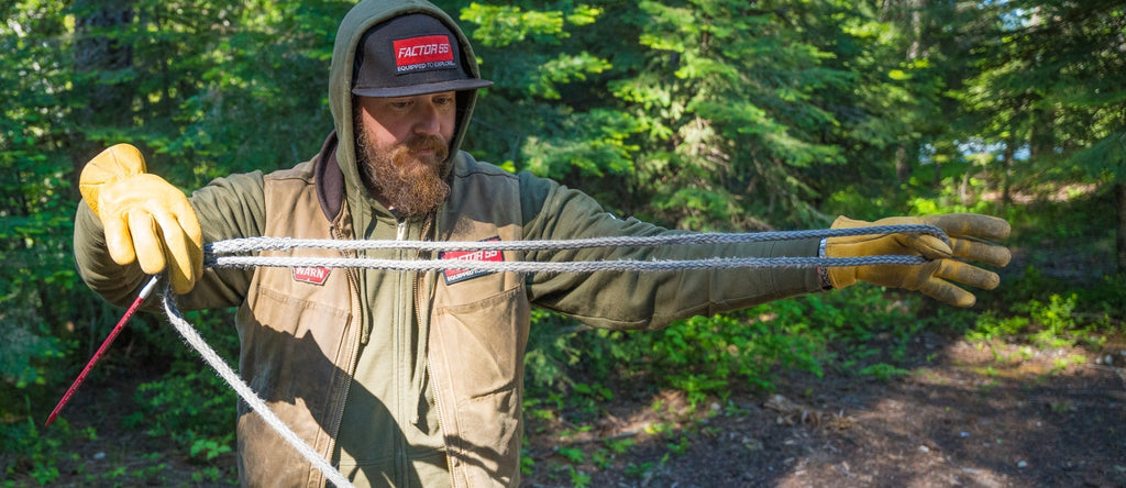 Bearded man in forest, wearing gloves and a cap, carefully inspecting winch rope for repair