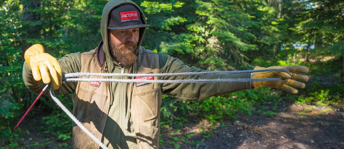 Bearded man in forest, wearing gloves and a cap, carefully inspecting winch rope for repair