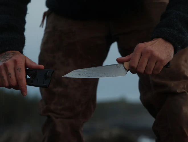 Person in brown pants holds a wet knife and black sheath, with tattoos visible on the left hand.