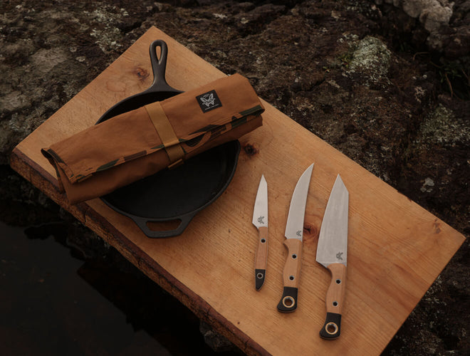 Three wood-handled knives, a cast iron skillet, and a tan canvas roll on a wooden board over dark rocks.