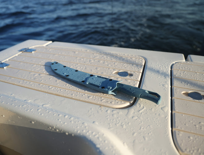 A teal knife in a sheath lies on the wet, white deck of a boat, with dark blue water in the background.