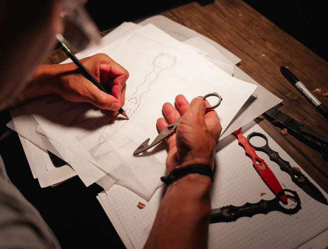 Person sketches a knife design with pencil, holding metal prototype; red and black finished knives rest on graph paper nearby.