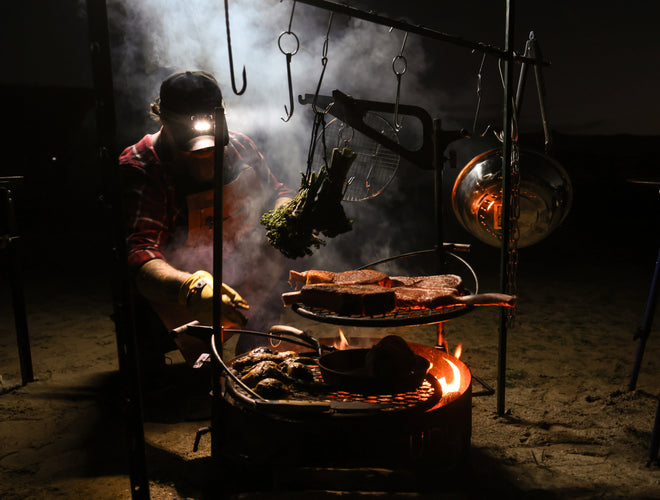 Man in plaid shirt and headlamp grills steaks over smoky campfire, hanging greens nearby.